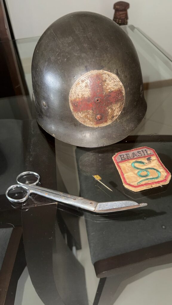 alt="cidadania italiana por descendência — pracinha Antonio Pieri Junior FEB. Objetos originais de Antonio Pieri Junior: capacete de enfermeiro de campanha com cruz vermelha, tesoura cirúrgica e patch da FEB. Arquivo familiar."